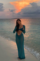 Woman in a teal dress standing on a beach at sunset.