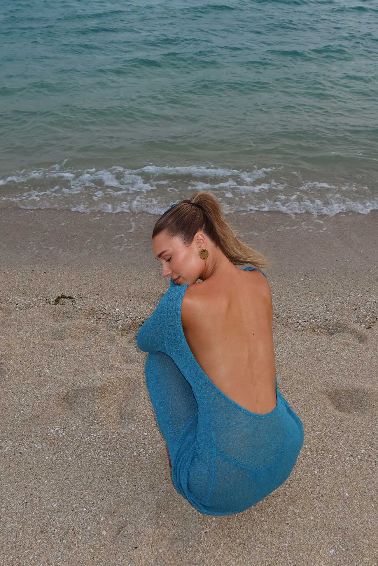 Woman in a teal dress sitting on a beach with ocean waves in the background
