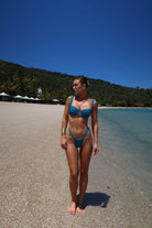 Woman in a teal bikini standing on a sandy beach with clear blue water and sky.