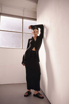 Woman in a black outfit standing against a white wall with a window in the background