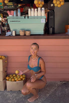 Woman in a teal bikini squatting next to a fruit stand with oranges.
