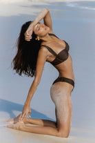 Woman in a chocolate brown bikini posing on a beach with clear blue sky.