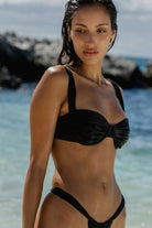 Woman in a black bikini standing on a beach with ocean and rocks in the background