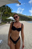 Woman in a black bikini standing on a beach with palm trees and mountains in the background.