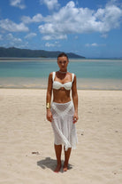 Woman in a white bikini top and skirt standing on a sandy beach with clear blue water and mountains in the background.