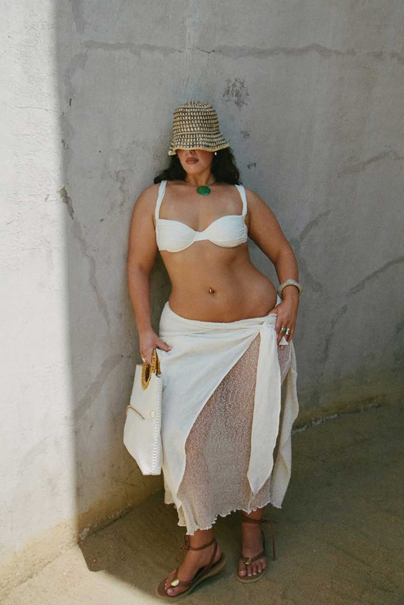 Woman wearing a white bikini top and skirt with a straw hat against a plain wall.