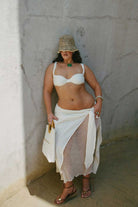 Woman wearing a white bikini top and skirt with a straw hat against a plain wall.