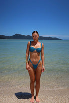 Woman in a teal bikini standing on a beach with clear water and mountains in the background