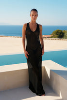 A woman stands outdoors by a pool wearing a sheer black sleeveless maxi dress. The background features a clear blue sky, ocean, and white architectural structures.