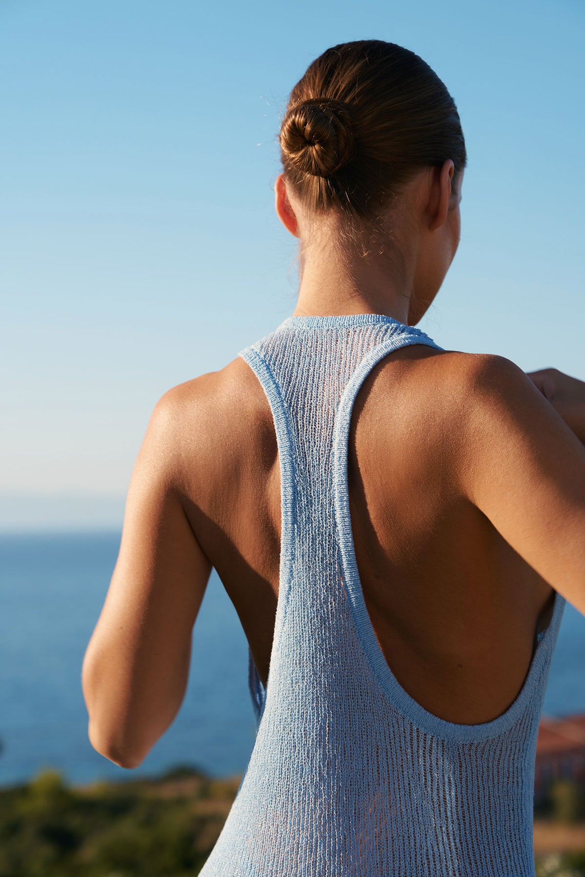 A woman viewed from behind wearing a light blue ribbed knit tank top, standing outdoors with the ocean and blue sky in the background.