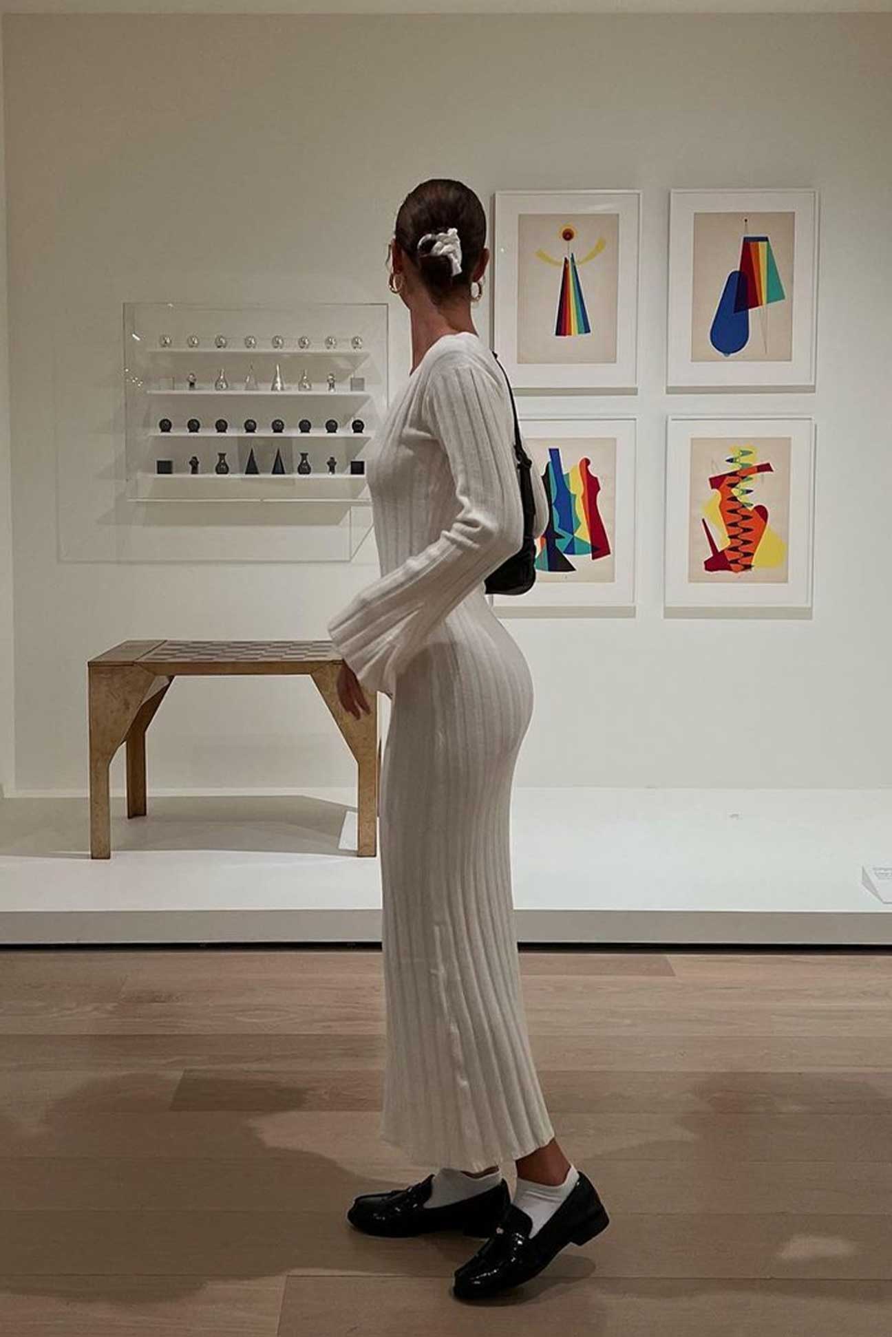 Woman in a white dress standing in an art gallery with colorful abstract paintings on the wall.