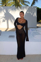 Person wearing a black sheer dress in an outdoor setting with palm trees and a white wall.