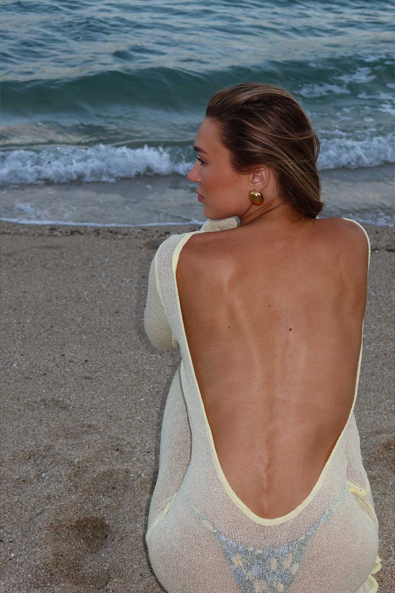 Woman in a yellow dress with a deep backless design sitting on a beach.