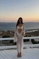 Woman in a white outfit standing on a balcony with a scenic view of the sea and sunset.