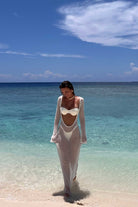 Woman in a white dress standing on a beach with clear blue water and sky.