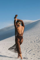Person in a sheer outfit standing on a sandy dune with a clear blue sky.