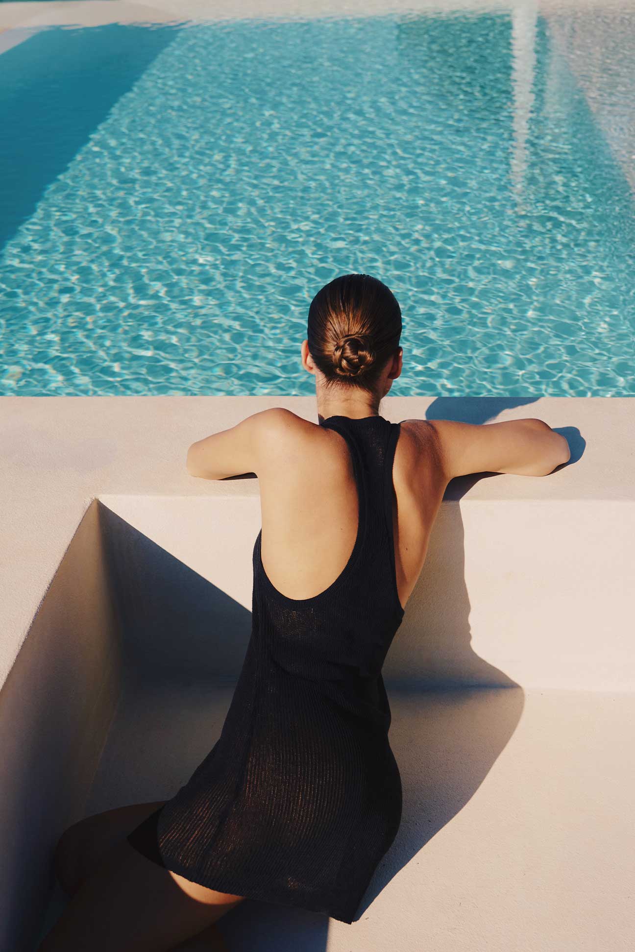 A woman seen from behind wearing a black mesh backless dress, leaning on the edge of a white concrete pool structure with bright blue water in the background.