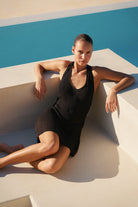 A woman reclining on a white concrete poolside step wearing a black ribbed knit halter mini dress.