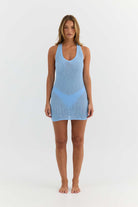 A woman stands barefoot against a plain white studio background wearing a light blue sleeveless knit mini dress with a scoop neckline and a V-shaped panel detail at the waist.