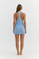 A woman viewed from the back wearing a light blue sleeveless mini dress with a racerback design and a sheer mesh overlay, set against a plain white studio background.