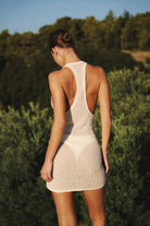 A woman viewed from behind wearing a sheer, open-back mini dress in a light cream or off-white color, set against a natural background of green trees and bushes.