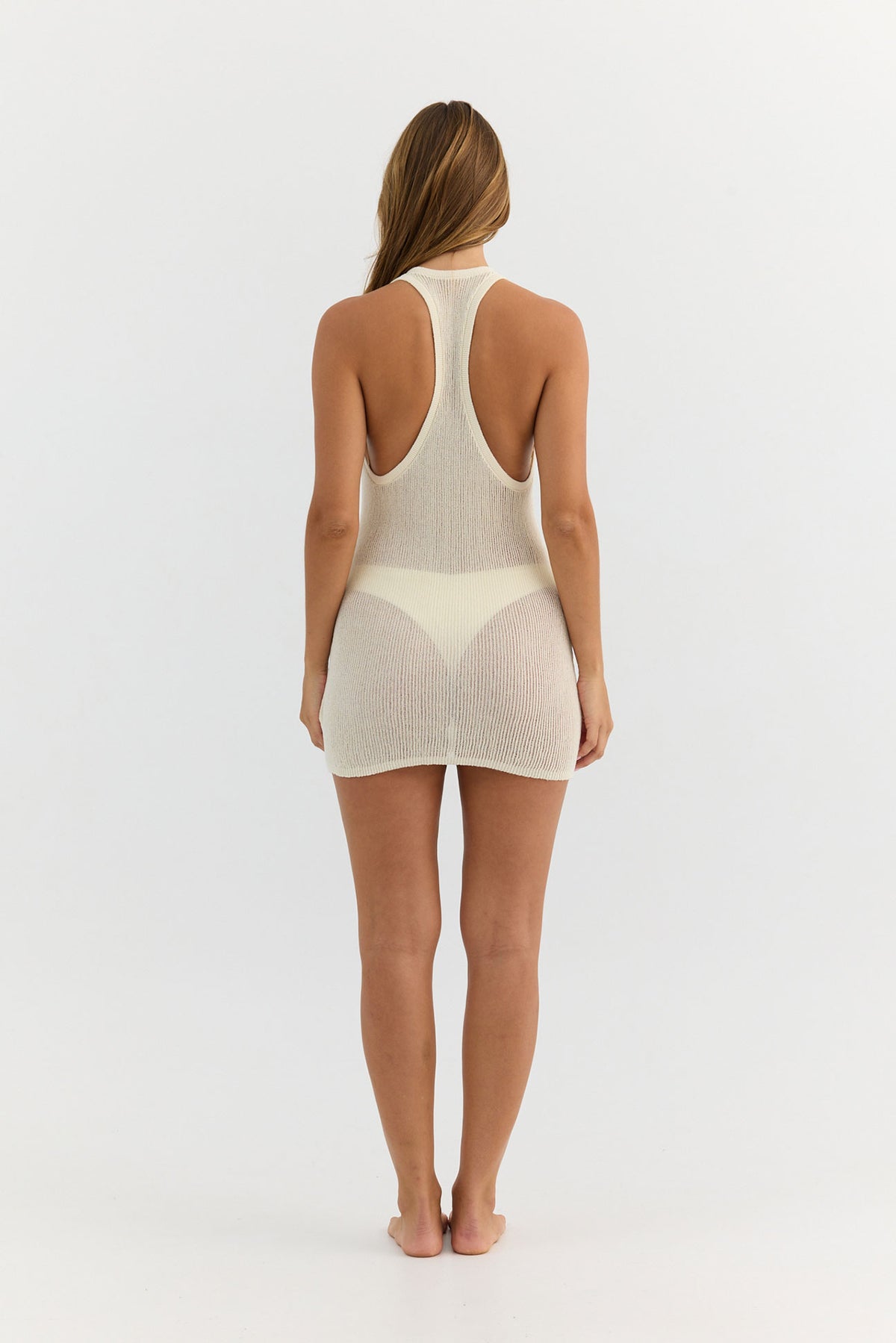 A woman viewed from the back wearing a light cream-colored, semi-sheer knit one-piece bodycon mini dress with a racerback design and a high-cut thong-style bottom.