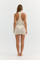 A woman viewed from the back wearing a light cream-colored, semi-sheer knit one-piece bodycon mini dress with a racerback design and a high-cut thong-style bottom.