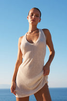 A woman wearing a ribbed knit sleeveless mini dress in a light cream color, standing outdoors with the blue ocean and sky in the background.