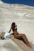 Person in a brown bikini sitting on a towel with a salt block and towel beside them, against a white sand dune background.