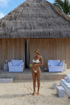 Woman in a white bikini standing on a sandy beach in front of a thatched-roof hut with outdoor furniture.