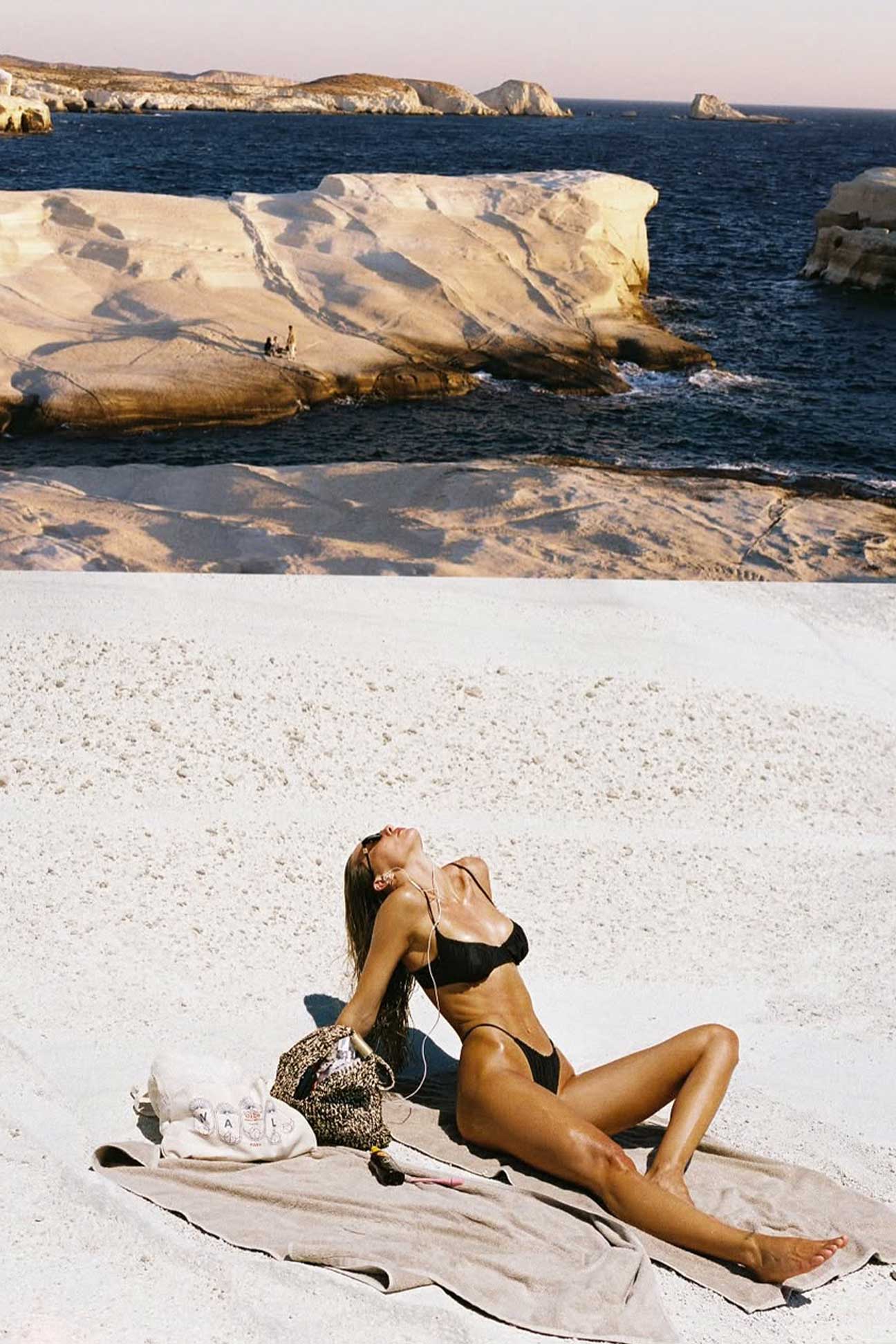 Woman in a brown bikini lying on a towel by the ocean with cliffs in the background
