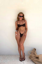 Woman wearing a brown bikini against a white wall with a straw bag on the floor.