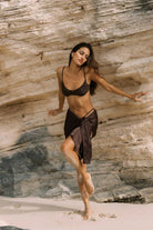 Woman in a black bikini and sarong standing on a rocky beach.