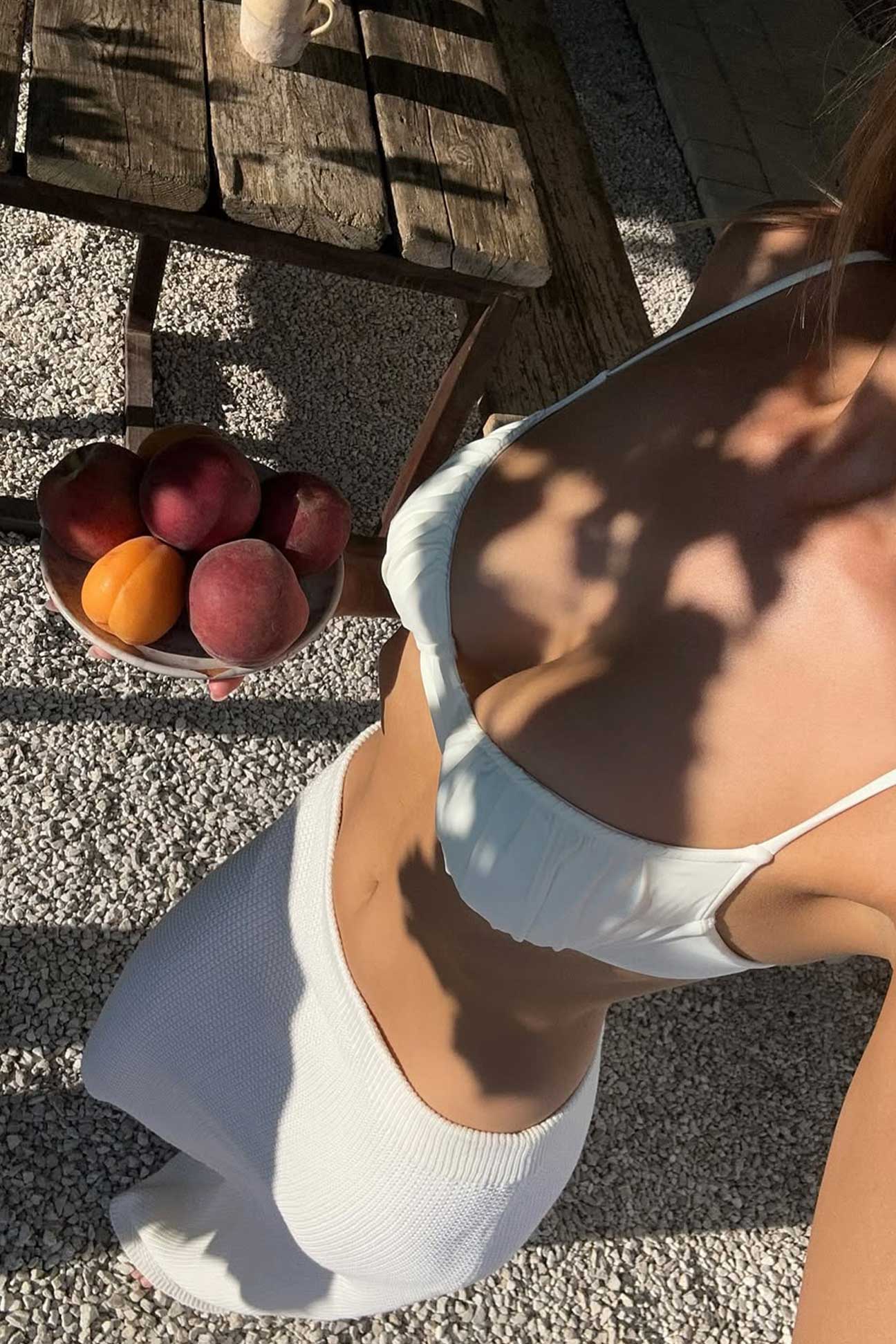 Person wearing a white tank top sitting on a wooden bench with peaches in a bowl.