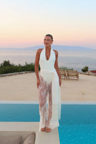 A woman stands by a swimming pool at sunset wearing a white sleeveless halter dress with sheer, patterned white and beige tulle layers. The background features a calm sea, distant mountains, and a beige terrace with lounge chairs.