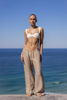 A model stands outdoors by the ocean wearing loose beige wide-leg pants and a white bralette, photographed against a clear blue sky and sea background.