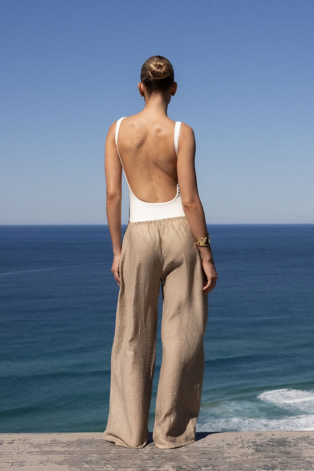 A woman stands with her back to the camera, looking out over a blue ocean under a clear sky. She is wearing a white open-back one-piece swimsuit and beige wide-leg linen trousers.