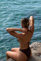 Woman in a brown bikini sitting by the water with her hair tied back.