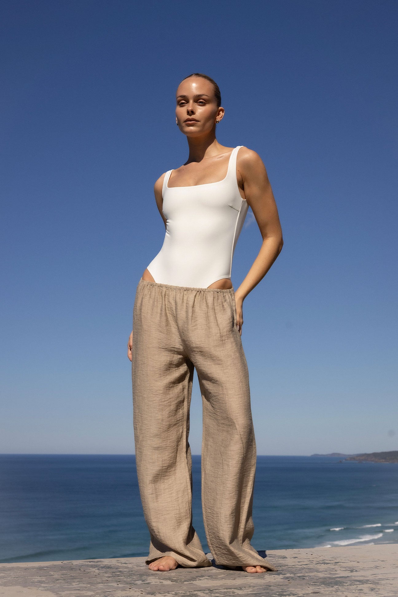 A woman stands outdoors by the ocean wearing a fitted white bodysuit and loose beige linen pants against a clear blue sky.
