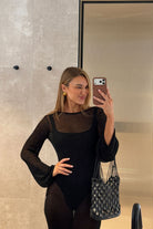 Woman taking a mirror selfie wearing a black outfit with sheer sleeves.