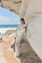 Woman in a white outfit leaning against a rock formation by the ocean.