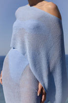 A close-up of a model wearing a light blue, semi-sheer knit shawl or wrap draped over her shoulders and torso, with a blue sky and ocean in the background.