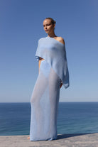 A woman stands outdoors by the sea wearing a flowing, semi-sheer light blue wrap dress or cover-up with draped sleeves against a clear blue sky.