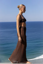 A woman stands in profile on a beach wearing a dark brown two-piece set consisting of a cropped bandeau top and a matching long skirt. The background features a clear blue sky and the ocean.