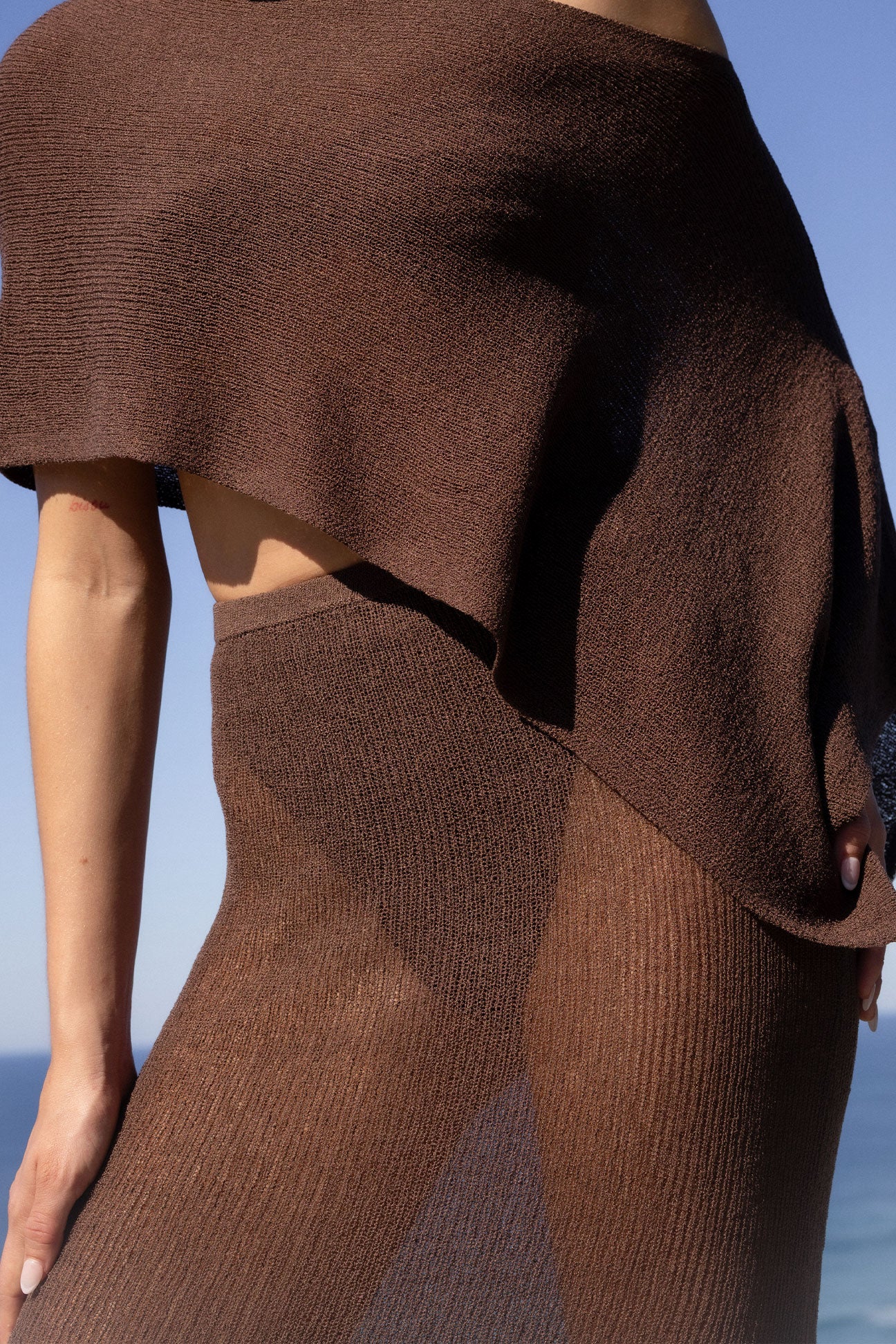 A close-up of a model wearing a brown ribbed knit poncho-style top and matching skirt against a blue sky and sea background.