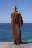 A model stands barefoot on a stone ledge overlooking the ocean, wrapped in a draped, one-shoulder brown knit cover-up or dress.