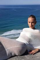 A woman reclines on a stone ledge by the ocean, wrapped in a sheer white knit shawl or poncho. The background features a calm blue sea under a clear sky.