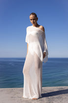 A woman stands barefoot on a stone ledge overlooking the ocean, wearing a sheer, off-the-shoulder white maxi dress or cover-up against a clear blue sky and sea background.