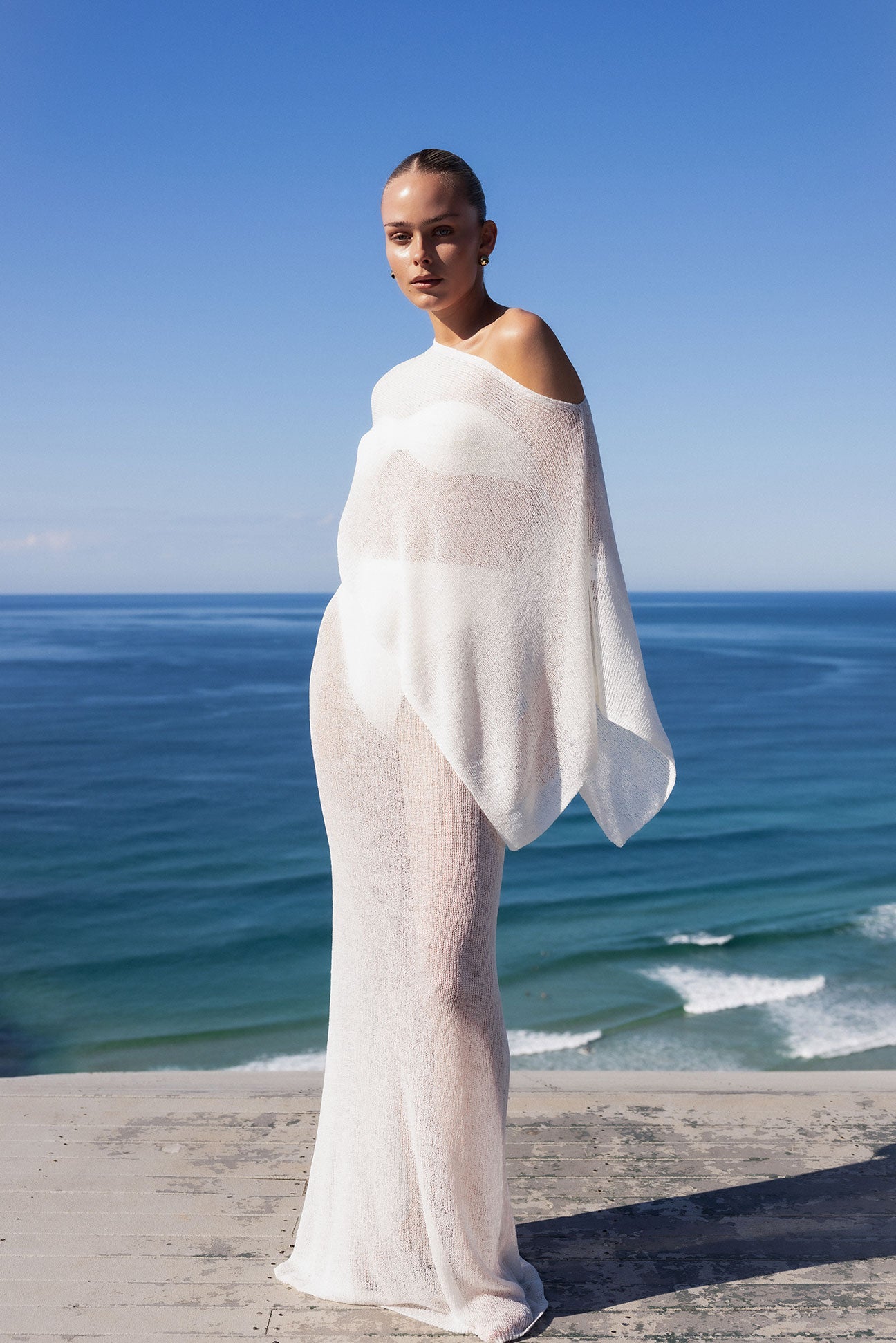 A woman stands on a beach wearing a sheer, off-the-shoulder white maxi dress or tunic with a flowing cape-like drape. The background features a clear blue sky and ocean.