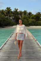 Woman in a white top and silver skirt standing on a wooden pier with turquoise water and greenery in the background.